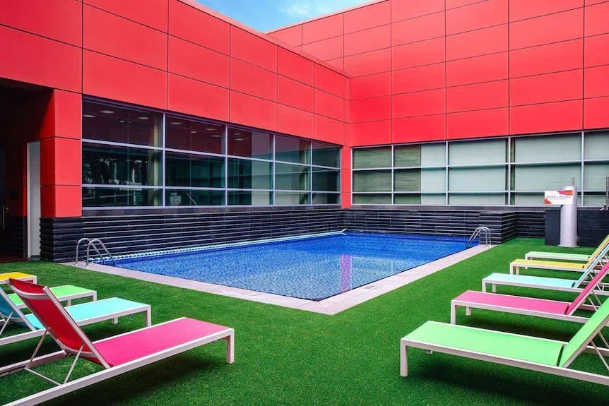 Modern outdoor swimming pool with colorful lounge chairs on green turf, next to a red building.