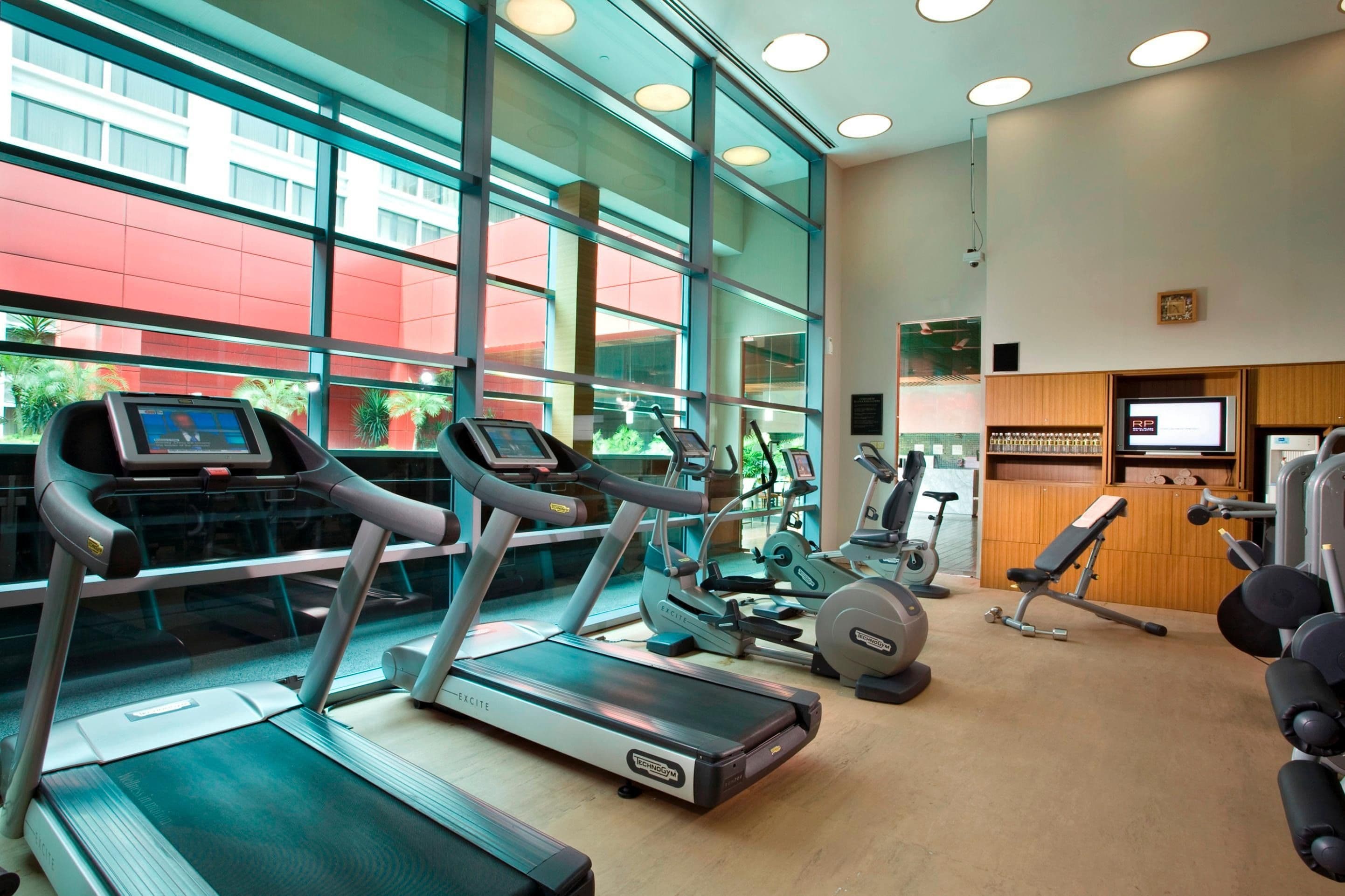 Modern gym with treadmills, elliptical machines, and large windows overlooking city buildings.
